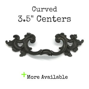 May include: A dark, ornate metal drawer pull with a curved design and intricate scrollwork details. The text above the handle reads Curved 3.5" Centers, and the text below includes a green plus sign followed by the words More Available.