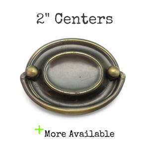 May include: Antique brass-toned drawer pull with an oval center and a curved handle. The pull has a 5.08 cm center-to-center measurement. The text "2" Centers" is above the pull, and "More Available" is below.