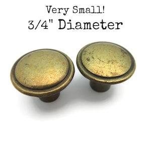 May include: Two small, antique gold-toned cabinet knobs, each with a 3/4 inch diameter.  "Very Small! 3/4" Diameter" is written in black text above the knobs.
