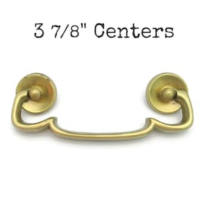 May include: A brass-toned drawer pull with a curved handle and circular end pieces. The text "3 7/8" Centers" is printed above the pull. The pull is designed for furniture and has a vintage aesthetic.