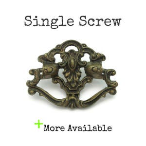 May include: Ornate antique brass drawer pull with a decorative design. The pull features a central oval element and curved handles. The text "Single Screw" is above the pull, and "More Available" is below it.