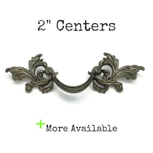 May include: An antique bronze-toned metal drawer pull featuring ornate, symmetrical scrollwork and leaf-like detailing. The handle has a curved centre bar with a dotted texture. Text above reads 2" Centers and below reads + More Available.