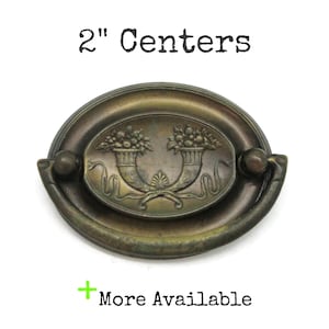 May include: An oval-shaped brass drawer pull with a dark, aged patina. The center plate features an embossed design of two cornucopias filled with fruit and foliage. A swinging bail handle is attached to the sides. Text reads 2" Centers.