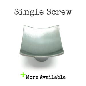 May include: A single, square-shaped cabinet knob with a brushed metal finish. The knob has a slightly curved top and a rounded base. The text "Single Screw" is above the knob, and "More Available" is below it.
