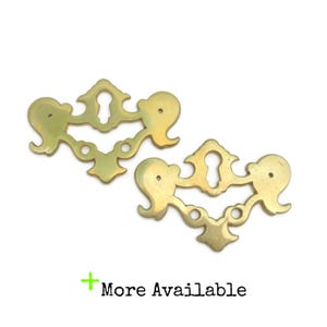 May include: Two brass-colored decorative drawer pulls with a keyhole design. Each pull has a symmetrical, ornate shape with curved edges and small holes. The text "More Available" is in green.