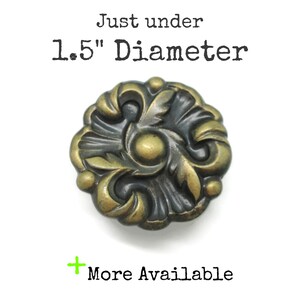 May include: A round, antique brass-colored metal knob featuring a raised floral relief pattern with a central circular detail. The text "Just under 1.5" Diameter" and "+ More Available" is printed above and below the object respectively.