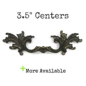 May include: An ornate, dark bronze-toned metal drawer pull featuring intricate scrollwork and leaf-like detailing. The text above reads 3.5" Centers, and the text below includes a green plus sign followed by the words More Available.