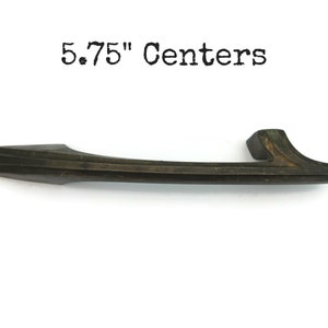 May include: A dark brown metal cabinet pull with a 5.75" center to center measurement. The pull has a simple, modern design with a slightly curved shape.
