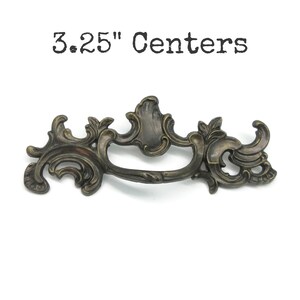 May include: An ornate, dark bronze-toned metal drawer pull with intricate scrollwork and leaf-like detailing. The hardware features a central handle with a curved design and includes the text 3.25" Centers printed above the object.