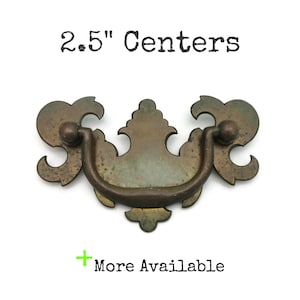 May include: Antique brass drawer pull with a decorative design. The pull has a curved handle and ornate details, with a 2.5-inch center-to-center measurement. The text "2.5" Centers" is above the pull, and "More Available" is below.