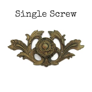 May include: An antique brass-toned furniture pull with an ornate, symmetrical design. The pull features a central circular element surrounded by stylized floral and leaf motifs. The text "Single Screw" is above the pull.