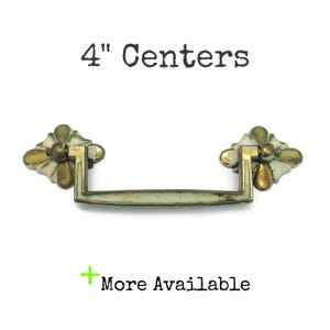 May include: A metal drawer pull with a brass and off-white finish, featuring a drop handle and two decorative mounting plates. The text 4" Centers is displayed above the handle, and the text + More Available is displayed below the handle.
