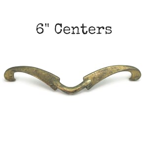 May include: A vintage-style brass drawer pull with a curved design. The pull has a distressed finish and the text "6" Centers" printed above it. The pull measures 6 inches between the centers of the mounting holes.