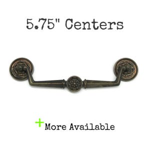 May include: A bronze-toned metal drawer pull with a swinging handle, featuring circular rosettes at the ends and a central decorative medallion. The text 5.75" Centers and + More Available is printed above and below the hardware piece.