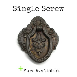 May include: A dark bronze-toned metal drawer pull with a shield-shaped backplate and a swinging drop handle. The piece features embossed scrollwork and floral details. Text at the top reads Single Screw and at the bottom reads + More Available.