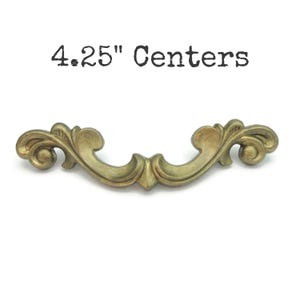 May include: Ornate brass-toned drawer pull with a curved design. The pull features a decorative, scroll-like pattern and the text "4.25" Centers" in black. The hardware is designed for furniture and has a vintage aesthetic.