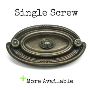 May include: An oval-shaped metal drawer pull with an aged bronze finish, featuring a rope-style border and a central decorative medallion. The text "Single Screw" appears above the hardware, and "+ More Available" is printed below.