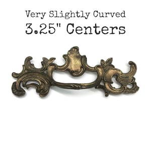 May include: A bronze-toned metal drawer pull with an ornate, scrolling design and a central handle. The text above the object reads: Very Slightly Curved 3.25" Centers. The hardware features a weathered finish with dark highlights and intricate curves.