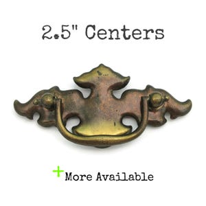 May include: An antique brass cabinet pull with a decorative, ornate design. The pull has a D-shaped handle and is 2.5" between the screw holes.
