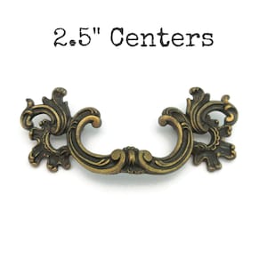 May include: An antique-style bronze drawer pull with intricate scroll detailing and a curved handle. The text "2.5" Centers" is printed above the pull. The pull measures 6.35 cm between the screw holes, a classic hardware piece.