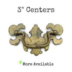 May include: A vintage-style brass drawer pull with a curved handle and ornate floral design. The pull measures 3 inches between the centers of the mounting holes. The text "3" Centers" is above the pull, and "More Available" is below.