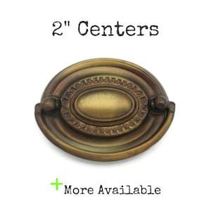 May include: Oval-shaped antique brass drawer pull with a 2-inch center-to-center measurement. The pull features an ornate, detailed design with a central oval and decorative edging. The text "2" Centers" and "More Available" are also visible.