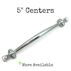 May include: A polished silver cabinet pull with a 5-inch center-to-center measurement. The handle has a curved design with decorative ends. The text "5" Centers" is above the handle. The text "More Available" is below the handle.