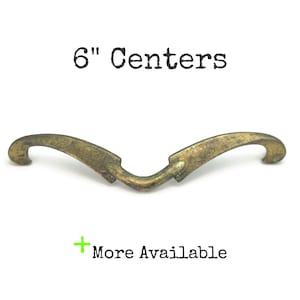 May include: Antique brass-toned cabinet pull with a curved design. The pull measures 6 inches (15.24 cm) between the centers of the mounting holes. The text "6" Centers" is above the pull, and "More Available" is below.