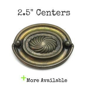 May include: An oval-shaped brass drawer pull with a dark, aged patina and a central swirling sunburst pattern. The hardware features a drop handle and is labeled with the text 2.5" Centers and More Available.