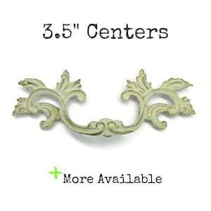 May include: A decorative drawer pull with a light green finish and gold-toned highlights, featuring an ornate, scrolling leaf design. The text 3.5" Centers is displayed above the pull, and the text + More Available is displayed below.
