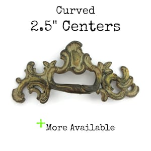 May include: Ornate, antique-style drawer pull with a curved design. The pull is approximately 2.5 inches (6.35 cm) between centers, with a bronze and gold finish. The text "Curved 2.5" Centers" is visible.