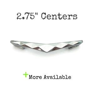 May include: A silver-toned cabinet pull with a faceted, V-shaped design. The text "2.75" Centers" is above the pull, and "More Available" is below. The hardware is designed for furniture and cabinetry.