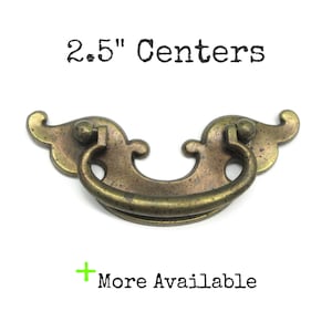 May include: A brass-toned metal drawer pull with a curved, ornate backplate and a swinging bail handle. The text 2.5" Centers is printed above the hardware, and a green plus sign with the text More Available is printed below the hardware.
