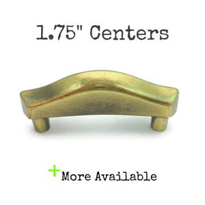 May include: A brass-colored cabinet pull with a curved design. The pull has two small legs and the text "1.75" Centers" above it. The text "More Available" is below the pull.