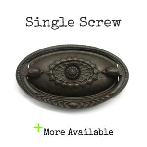 May include: An oval-shaped dark bronze drawer pull with a central floral medallion and a swinging handle. The text "Single Screw" appears above the hardware, and the text "+ More Available" is positioned below the object on a white background.
