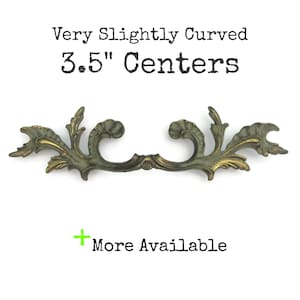May include: A decorative, slightly curved brass drawer pull with a floral design. The pull measures 3.5 inches (8.89 cm) between the centers of the mounting holes. The text "More Available" is also present.