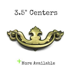 May include: A brass drawer pull with a curved, ornate design and a central decorative element. The hardware features two mounting points with 3.5" centers. The text "3.5" Centers" and "+ More Available" is printed above and below the object.