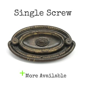 May include: An oval-shaped bronze-toned metal drawer pull with a weathered finish, featuring a central decorative medallion and a rope-style border. The text "Single Screw" appears above the object, and "+ More Available" is printed below.