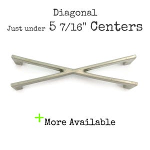 Peut inclure: Une poignée de meuble diagonale argentée avec un design en forme de croix. Le texte indique "Diagonal Just under 14.13 cm Centers" et "More Available". Le matériel est de style moderne.