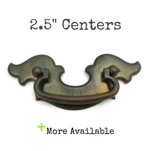 May include: An antique brass cabinet pull with a decorative wing design. The pull has a distressed finish and a brown metal bar handle. It is 2.5" between centres.