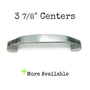 May include: Silver cabinet hardware pull with a 9.8 cm centre to centre measurement. The pull has a curved shape with a decorative design on the ends.