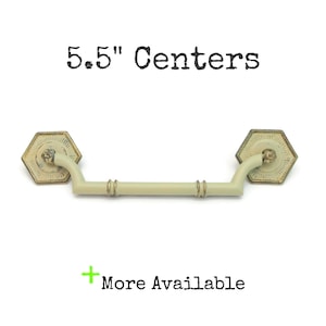 May include: Cream-colored cabinet pull with a 5.5-inch center-to-center measurement. The pull has a hexagonal backplate with an aged gold finish and a curved handle. The text "5.5" Centers" is above the pull, and "More Available" is below.