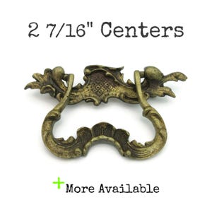May include: Ornate antique brass drawer pull with a curved handle and decorative details. The text "2 7/16" Centers" is above the pull. The text "More Available" is below the pull.