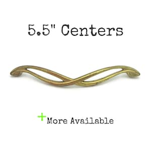 May include: A vintage-style brass cabinet pull with a wavy design. The text "5.5" Centers" is above the pull, and "More Available" is below. The pull is approximately 5.5 inches long.