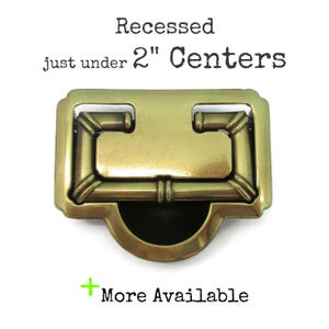 May include: A brass-toned metal recessed drawer pull with a rectangular frame and a central hinged handle. The text above reads Recessed just under 2" Centers and the text below reads + More Available. The hardware has a smooth, metallic finish.