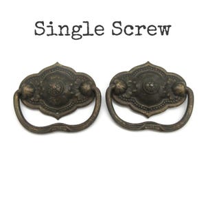 May include: Two antique bronze-toned metal drawer pulls with a decorative backplate and a swinging handle. The text "Single Screw" is printed in black above the hardware. Each pull features a central circular motif with floral details.