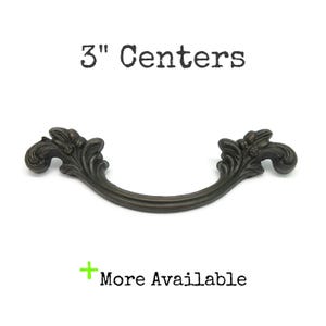 May include: A dark bronze-toned metal drawer pull with ornate, curved leaf-like detailing on both ends. The handle features a central arched bar and is labelled with the text 3" Centers and More Available, indicating the mounting distance.