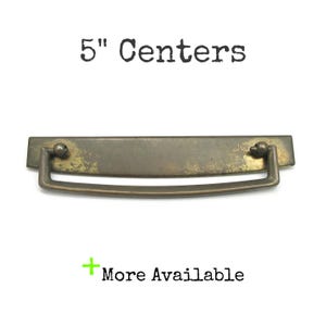 May include: A brass-toned metal drawer pull with a rectangular backplate and a swinging bail handle. The hardware features a weathered, aged finish. Text above reads 5" Centers and text below reads + More Available.