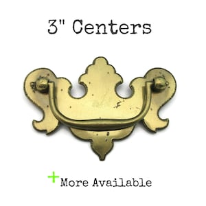 May include: A brass-toned drawer pull with a decorative design. The pull has a curved handle and ornate backplates. The text "3" Centers" is above the pull, and "More Available" is below it.