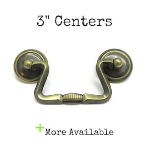 May include: A brass-toned metal drawer pull with a swinging handle and two circular mounting plates. The hardware features a textured center grip and is labeled with the text 3" Centers and + More Available.